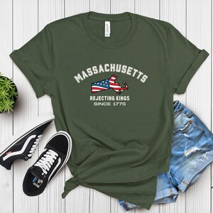 May include: Olive green t-shirt featuring the word "Massachusetts" above a graphic of the state's outline with an American flag design. The text "Rejecting Kings Since 1775" is below. The shirt is paired with denim shorts and black sneakers.