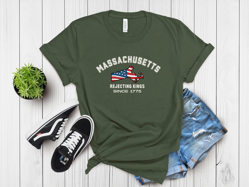 May include: Olive green t-shirt featuring the word "Massachusetts" above a graphic of the state's outline with an American flag design. The text "Rejecting Kings Since 1775" is below. The shirt is paired with denim shorts and black sneakers.
