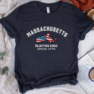 A navy blue t-shirt with a graphic design. The design features the word 'Massachusetts' in white arched text above a red, white, and blue graphic of the state's outline with the American flag. Below the graphic, the text reads 'Rejecting Kings Since 1775' in white.