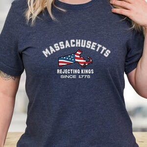 May include: Navy blue t-shirt with the word "Massachusetts" in white arched text above a graphic of the state's outline, incorporating the American flag. The text "Rejecting Kings Since 1775" is below the graphic.