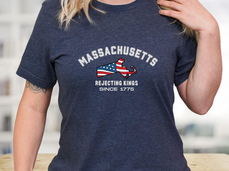 May include: Navy blue t-shirt with the word "Massachusetts" in white arched text above a graphic of the state's outline, incorporating the American flag. The text "Rejecting Kings Since 1775" is below the graphic.