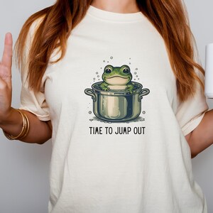 A cream-colored t-shirt featuring a cartoon frog in a pot. The frog is green with large eyes and is sitting in a silver pot. The text 'TIME TO JUMP OUT' is printed in black below the pot. The shirt is a casual style.
