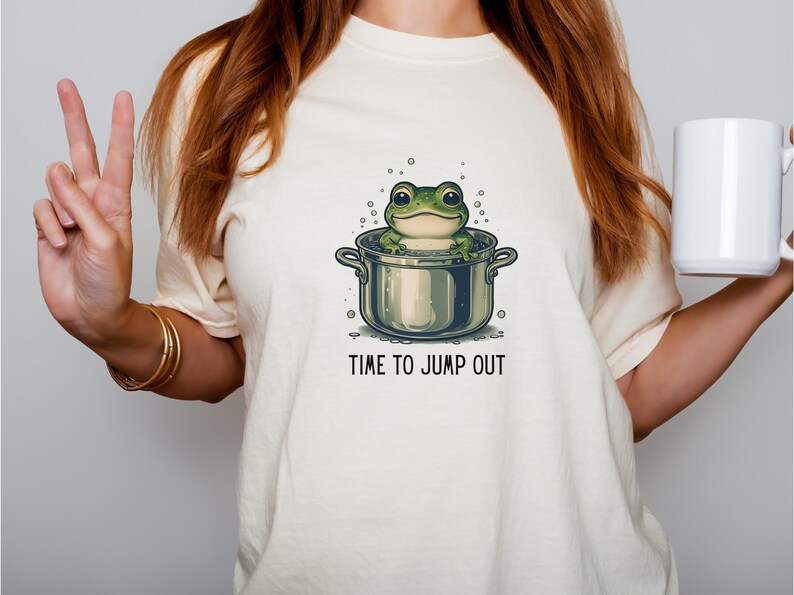 A cream-colored t-shirt featuring a cartoon frog in a pot. The frog is green with large eyes and is sitting in a silver pot. The text 'TIME TO JUMP OUT' is printed in black below the pot. The shirt is a casual style.