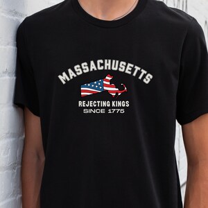 May include: Black t-shirt featuring the word "MASSACHUSETTS" in white arched text above a graphic of the state's outline with an American flag design. Below, the text reads "REJECTING KINGS SINCE 1775."