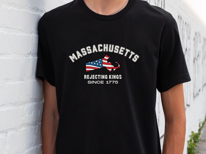 May include: Black t-shirt featuring the word "MASSACHUSETTS" in white arched text above a graphic of the state's outline with an American flag design. Below, the text reads "REJECTING KINGS SINCE 1775."