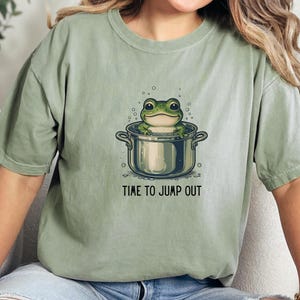 May include: A sage green short-sleeve t-shirt featuring a graphic of a green frog sitting inside a silver cooking pot with bubbles around it. Below the illustration, the text reads TIME TO JUMP OUT in a simple black sans-serif font.