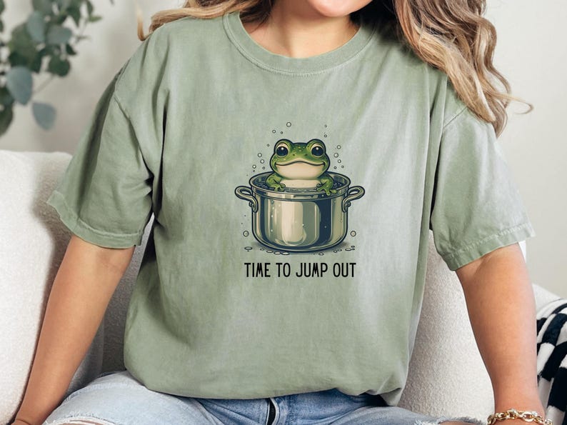 May include: A sage green short-sleeve t-shirt featuring a graphic of a green frog sitting inside a silver cooking pot with bubbles around it. Below the illustration, the text reads TIME TO JUMP OUT in a simple black sans-serif font.