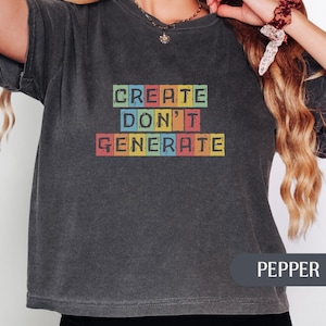 May include: A charcoal gray short-sleeve t-shirt featuring the text "CREATE DON'T GENERATE" printed in a block-letter style. Each letter is contained within a small, colorful square in shades of green, yellow, blue, and red, arranged in three rows.