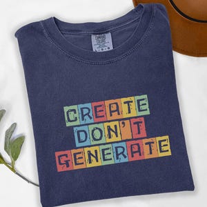 May include: Navy blue t-shirt with the text "CREATE DON'T GENERATE" in colorful block letters. The shirt is folded and has a crew neck. A brown hat is visible in the background.