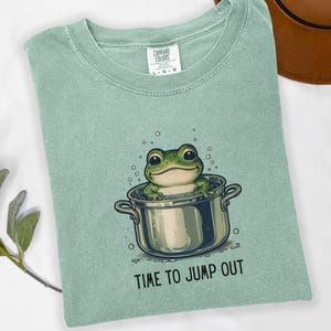 May include: A sage green t-shirt featuring a graphic of a green frog sitting inside a silver cooking pot with bubbles around it. Below the pot, the text reads "TIME TO JUMP OUT" in black capital letters. The shirt has a Comfort Colors brand label.