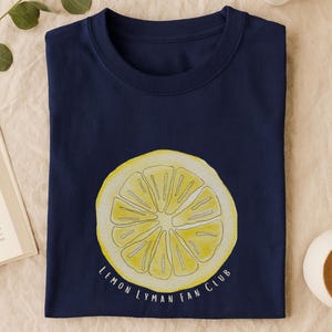 May include: Navy blue t-shirt featuring a watercolor illustration of a lemon slice. The text "Lemon Lyman Fan Club" is printed below the lemon. The shirt is folded neatly on a beige surface.