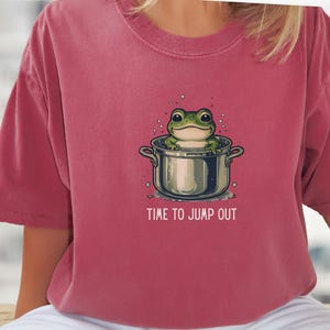 May include: A dusty rose-colored t-shirt featuring a graphic illustration of a green frog sitting inside a silver cooking pot. The text "TIME TO JUMP OUT" is printed in white capital letters below the pot, surrounded by small bubbles.