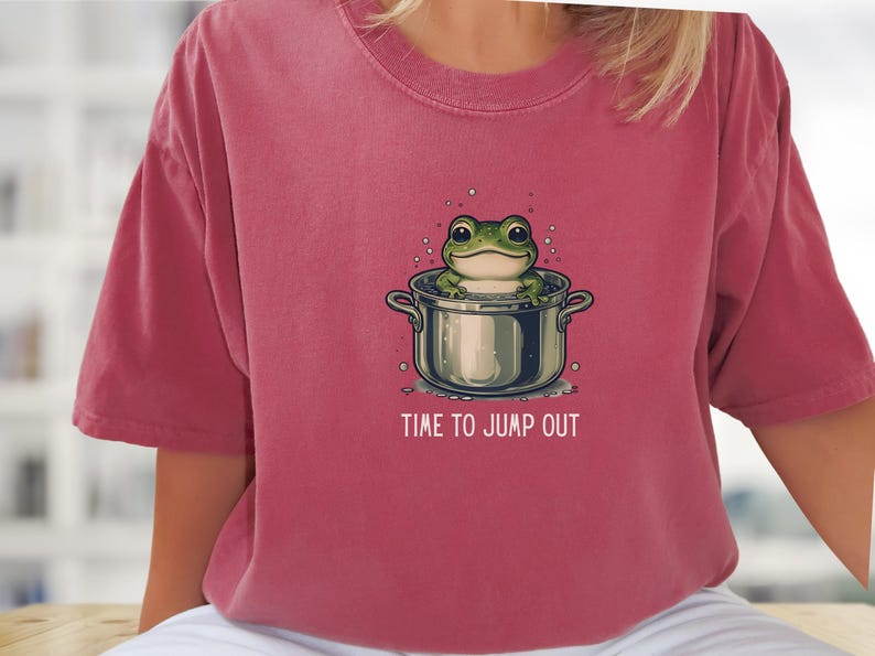 May include: A dusty rose-colored t-shirt featuring a graphic illustration of a green frog sitting inside a silver cooking pot. The text "TIME TO JUMP OUT" is printed in white capital letters below the pot, surrounded by small bubbles.