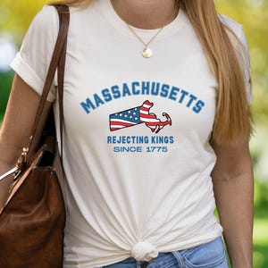 May include: Cream-colored t-shirt featuring the word "Massachusetts" in blue arched text above a graphic of the state's outline with a flag design. Below the graphic, the text reads "Rejecting Kings Since 1775."