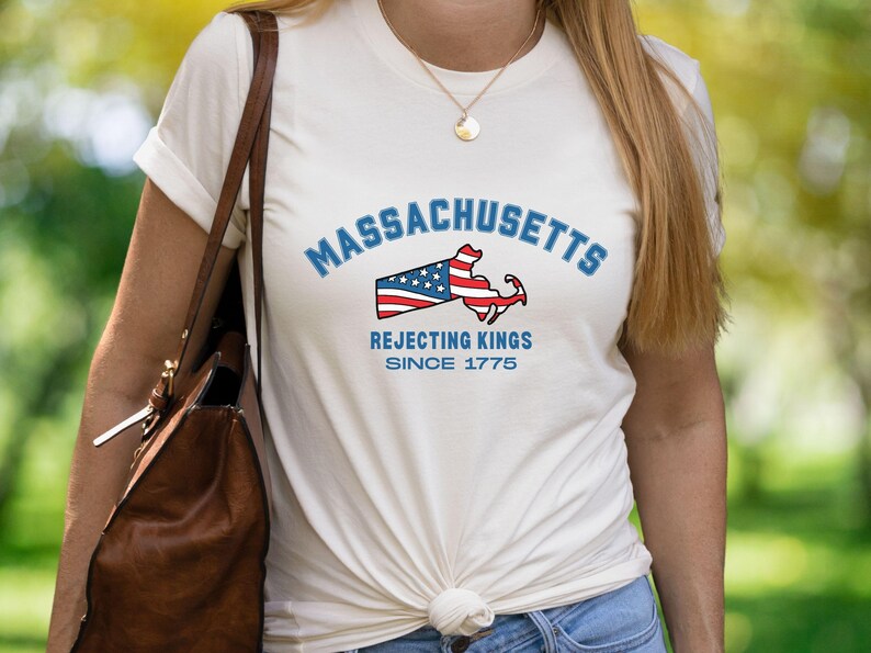 May include: Cream-colored t-shirt featuring the word "Massachusetts" in blue arched text above a graphic of the state's outline with a flag design. Below the graphic, the text reads "Rejecting Kings Since 1775."