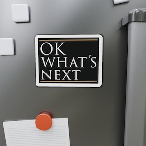 以下が含まれることがあります： 黒い背景に白い文字で「OK WHAT'S NEXT」と書かれた長方形のマグネット。細い金の縁で囲まれています。灰色の表面にあり、白い正方形のマグネットとオレンジ色の丸いマグネットが見えます。
