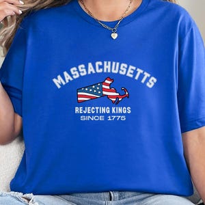 May include: Royal blue t-shirt featuring the word "Massachusetts" in white arched text above a graphic of the state's outline with a flag design. Below the graphic, the text reads "Rejecting Kings Since 1775".