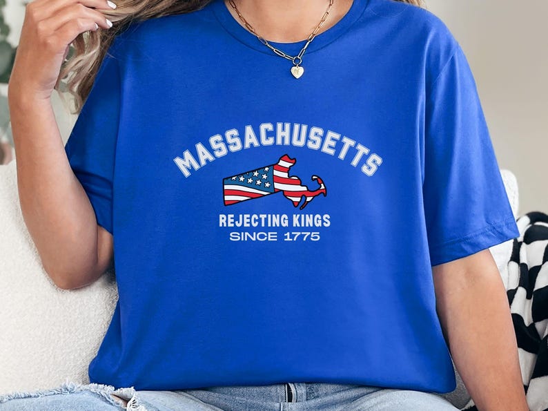 May include: Royal blue t-shirt featuring the word "Massachusetts" in white arched text above a graphic of the state's outline with a flag design. Below the graphic, the text reads "Rejecting Kings Since 1775".