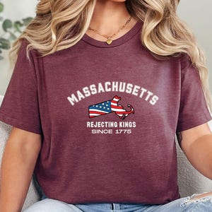 May include: Burgundy t-shirt featuring the word "Massachusetts" in white arched text above a graphic of the state's outline with an American flag design. Below the graphic, the text reads "Rejecting Kings Since 1775."