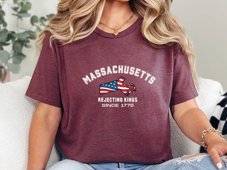 May include: Burgundy t-shirt featuring the word "Massachusetts" in white arched text above a graphic of the state's outline with an American flag design. Below the graphic, the text reads "Rejecting Kings Since 1775."