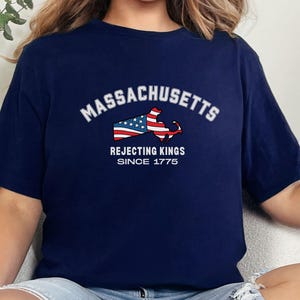 May include: Navy blue t-shirt featuring the word "Massachusetts" in white arched text above a graphic of the state's outline filled with the American flag. Below the graphic, the text reads "Rejecting Kings Since 1775."