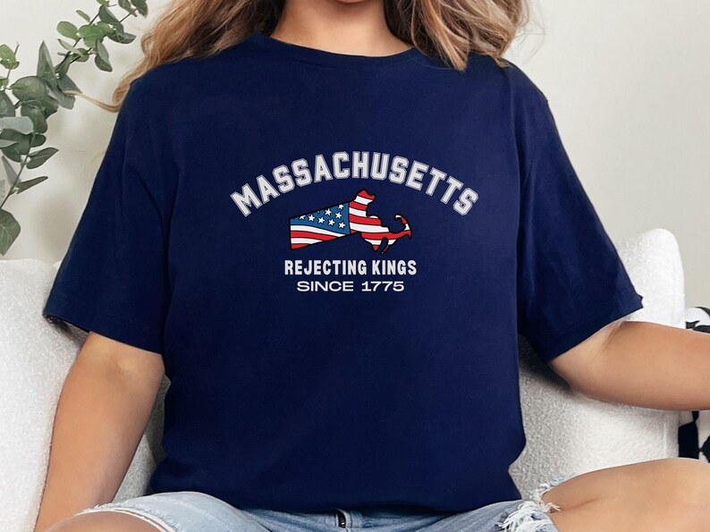 May include: Navy blue t-shirt featuring the word "Massachusetts" in white arched text above a graphic of the state's outline filled with the American flag. Below the graphic, the text reads "Rejecting Kings Since 1775."