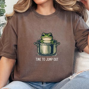 May include: A brown short-sleeve t-shirt featuring a graphic of a green frog sitting inside a silver cooking pot with bubbles around it. Below the pot, the text reads "TIME TO JUMP OUT" in white capital letters. The shirt has a crew neckline.