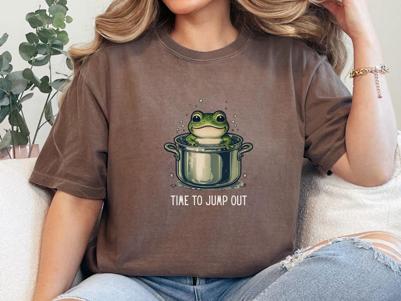 May include: A brown short-sleeve t-shirt featuring a graphic of a green frog sitting inside a silver cooking pot with bubbles around it. Below the pot, the text reads "TIME TO JUMP OUT" in white capital letters. The shirt has a crew neckline.