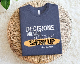 May include: A navy blue t-shirt with the quote "DECISIONS ARE MADE BY THOSE WHO SHOW UP" in white text, with an orange brushstroke underneath. The quote is attributed to Jed Bartlet. The shirt is displayed on a woven mat.