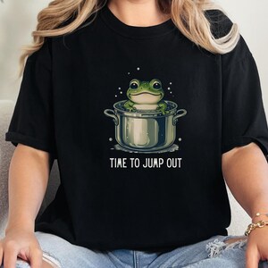 May include: A black t-shirt featuring a graphic of a green frog sitting inside a silver cooking pot. The text "TIME TO JUMP OUT" is printed in white capital letters below the pot. The design includes small white bubbles surrounding the frog.