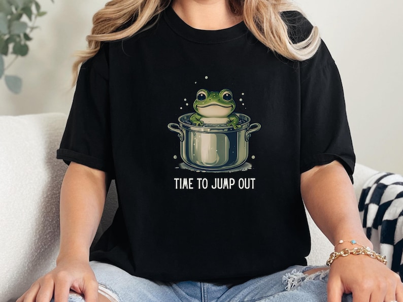 May include: A black t-shirt featuring a graphic of a green frog sitting inside a silver cooking pot. The text "TIME TO JUMP OUT" is printed in white capital letters below the pot. The design includes small white bubbles surrounding the frog.