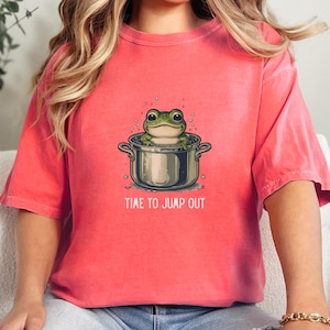 May include: A coral-colored short-sleeve t-shirt featuring a graphic of a green frog sitting inside a silver cooking pot. The text "TIME TO JUMP OUT" is printed in white capital letters below the pot, surrounded by small bubbles.