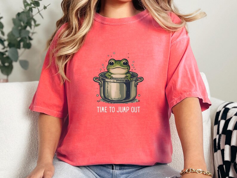 May include: A coral-colored short-sleeve t-shirt featuring a graphic of a green frog sitting inside a silver cooking pot. The text "TIME TO JUMP OUT" is printed in white capital letters below the pot, surrounded by small bubbles.