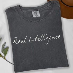 May include: A charcoal grey crew neck t-shirt with a faded, vintage-style wash. The shirt features the text "Real Intelligence" printed in a white, handwritten-style script across the chest. A Comfort Colors brand label is visible at the neckline.