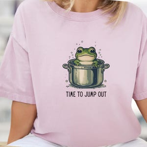 May include: A light pink short-sleeve t-shirt featuring a graphic of a green frog sitting inside a metal cooking pot with bubbles around it. Below the illustration, the text reads "TIME TO JUMP OUT" in a simple, dark, sans-serif font.