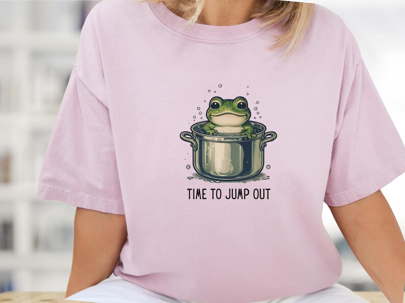 May include: A light pink short-sleeve t-shirt featuring a graphic of a green frog sitting inside a metal cooking pot with bubbles around it. Below the illustration, the text reads "TIME TO JUMP OUT" in a simple, dark, sans-serif font.