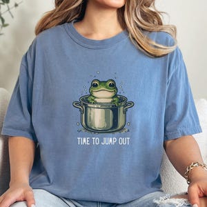 May include: A blue short-sleeve t-shirt featuring a graphic illustration of a green frog sitting inside a silver cooking pot with bubbles around it. Below the pot, the text reads TIME TO JUMP OUT in white capital letters centered on the chest.
