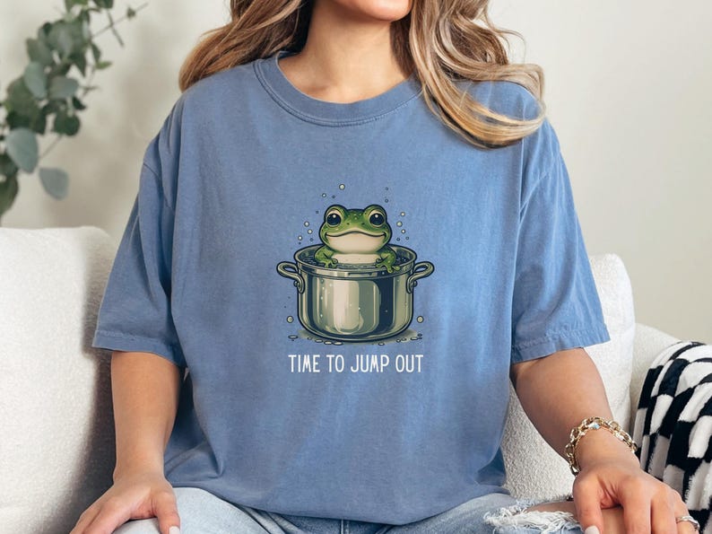 May include: A blue short-sleeve t-shirt featuring a graphic illustration of a green frog sitting inside a silver cooking pot with bubbles around it. Below the pot, the text reads TIME TO JUMP OUT in white capital letters centered on the chest.