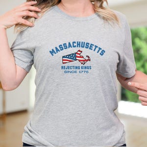 May include: A light gray t-shirt with the word "Massachusetts" in blue arched text above a graphic of the state's outline with an American flag design and an anchor. The text "Rejecting Kings Since 1775" is below the graphic.