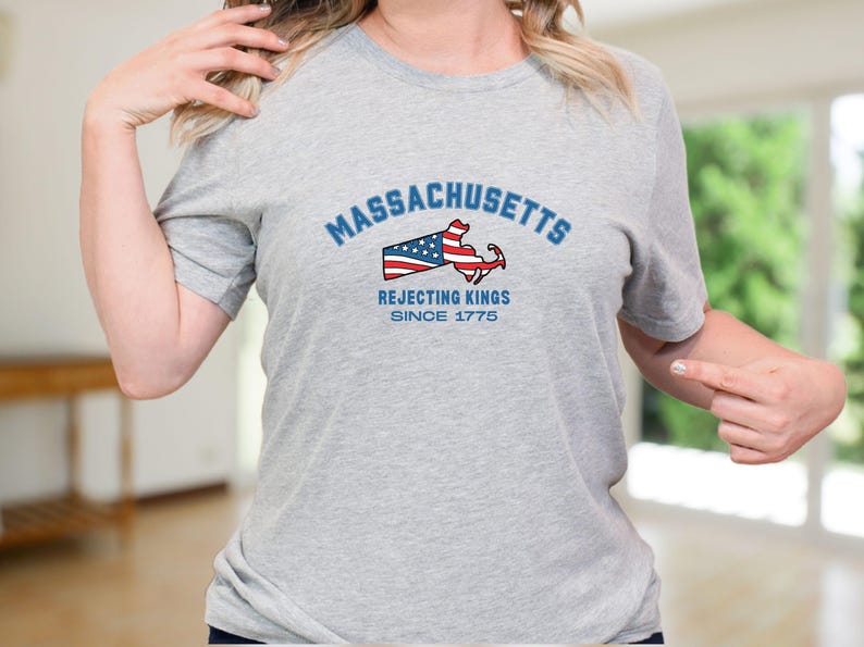May include: A light gray t-shirt with the word "Massachusetts" in blue arched text above a graphic of the state's outline with an American flag design and an anchor. The text "Rejecting Kings Since 1775" is below the graphic.