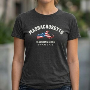 May include: Dark gray t-shirt featuring the word "Massachusetts" in white arched text above a graphic of the state's outline with an American flag design. Below the graphic, the text reads "Rejecting Kings Since 1775."