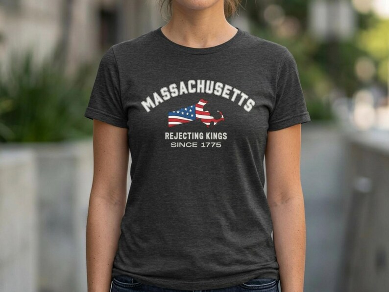 May include: Dark gray t-shirt featuring the word "Massachusetts" in white arched text above a graphic of the state's outline with an American flag design. Below the graphic, the text reads "Rejecting Kings Since 1775."