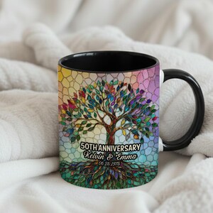 Könnte beinhalten: Eine schwarze Keramiktasse mit einem bunten Baumdesign im Buntglas-Stil. Der Text lautet 50TH ANNIVERSARY, Kelvin & Emma und 06.25.1975. Der Baum hat einen braunen Stamm und Wurzeln mit bunten Blättern vor einem pastellfarbenen Mosaikhintergrund.