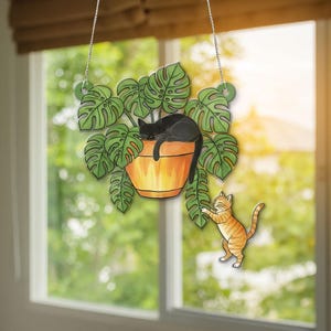 Könnte beinhalten: Dekoratives Hängeornament mit einer schwarzen Katze, die in einer Topfpflanze mit großen grünen Blättern schläft. Eine orangefarbene Tabbykatze greift nach der Pflanze. Das Ornament wird von einer silbernen Kette gehalten.