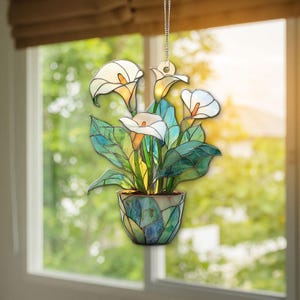 Könnte beinhalten: Glasmalerei-Kunstwerk, das Calla-Lilien in einem Topf darstellt. Die Blüten sind weiß mit orangefarbenen Zentren, die Blätter sind grün. Der Topf ist ein Mosaik aus blauem, grünem und beigem Glas. Eine Kette ist zum Aufhängen befestigt.