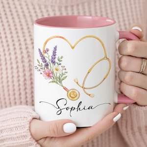 Könnte beinhalten: Ein weißer Keramikbecher mit rosafarbenem Innenraum und Henkel, verziert mit einem goldenen Stethoskop in Herzform. Ein Strauß aus violetten, rosa und weißen Blumen entspringt dem Stethoskopkopf. Der Name Sophia ist in schwarzer Schrift gedruckt.