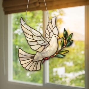 Puede incluir: Una paloma de vidriera con las alas extendidas, sosteniendo una rama de olivo. La paloma es blanca con acentos beige claro, y las alas están segmentadas. La rama de olivo tiene hojas verdes y acentos rojos. La paloma cuelga de una cadena plateada.