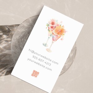May include: A white business card featuring a watercolour illustration of two martini glasses adorned with pink and orange flowers. The card displays the text "hi@yourwebsite.com, 925-657-4213, yourwebsite.com" and a QR code.