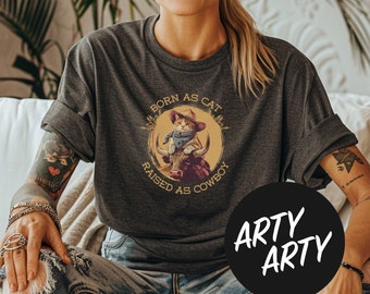 Pode incluir: Uma camiseta cinza com um gráfico de estilo vintage de um gato montando um touro. O gráfico diz "Born as Cat Raised as Cowboy".