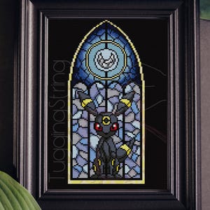 May include: A cross-stitch embroidery in a black frame depicting a black creature with red eyes and yellow rings in a stained-glass window style. The design features a blue circular moon motif above the creature, with the text TuggingString visible.